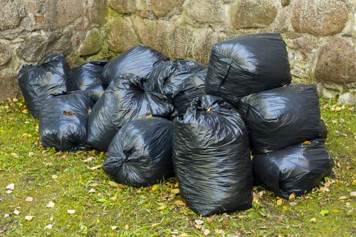 A group of approximately ten black plastic rubbish bags, filled with waste, are gathered on a patch of grass next to a stone wall. The bags appear tightly sealed, with some showing slight creases and folds on their surfaces, indicating they are filled with lightweight or soft materials. The setting is outdoor, with natural daylight illuminating the scene, highlighting the texture of the plastic and the surrounding green grass dotted with small fallen leaves. The stone wall behind the bags is constructed from irregularly shaped rocks with moss and lichen growth, suggesting an older boundary or garden feature. This image illustrates typical waste collection preparation in a residential or small commercial setting, matching the context of rubbish removal services such as those offered by Business Waste Removal Ealing, serving areas including Ealing within west London postcode regions.