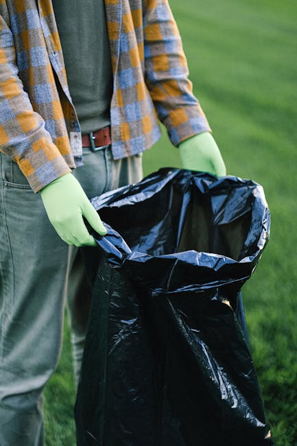 A person wearing a white glove is holding open a white polypropylene bag filled with garden waste, including small branches, leafy plant material, and scattered gravel or pebbles. In the background, t