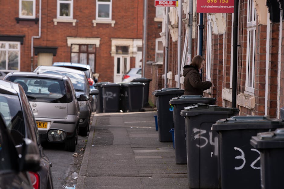 A row of four wheelie bins placed on the pavement beside a residential garden fence, with three black bins and one blue bin labeled with 'RMN'; the black bins have brown lids while the blue one has a 