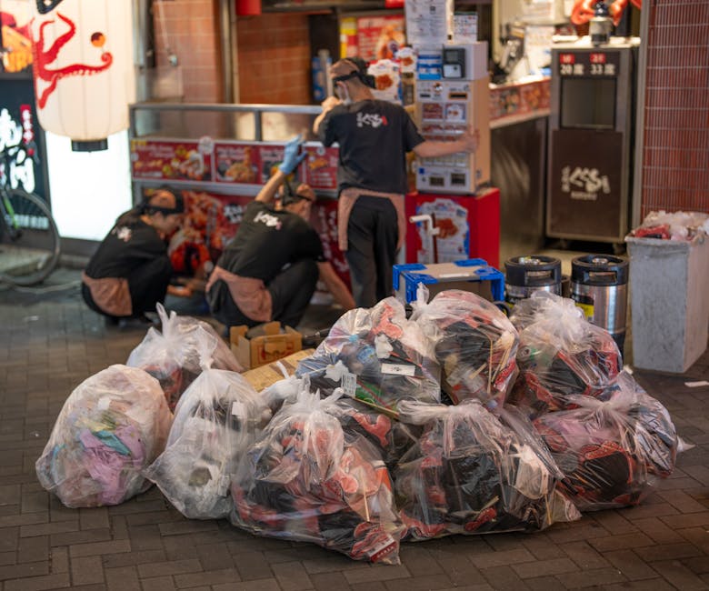 A person wearing a yellow and grey checkered jacket, grey trousers, and light green gloves is holding a large black rubbish bag open at the top, preparing for rubbish collection outdoors on a grassy a