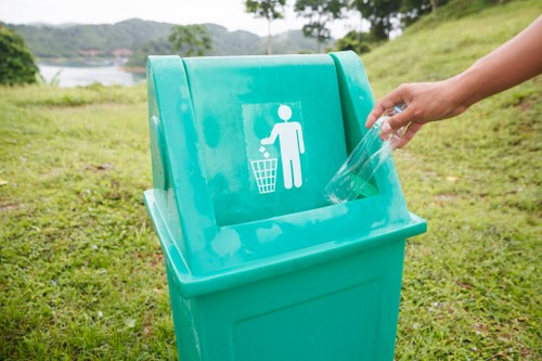 A person’s hand is seen placing a clear plastic bottle into a bright turquoise recycling bin situated outdoors on a grassy area. The bin features a white pictogram of a person disposing of waste into a trash can, indicating its use for recyclable plastics. In the background, there are trees, a gentle slope, and a body of water visible under a partly cloudy sky, suggesting a park or riverside setting. The scene is well-lit with natural daylight, and the focus is on the bin and the act of waste disposal, highlighting environmentally conscious rubbish management practices relevant to local outdoor spaces near Ealing and the surrounding areas.